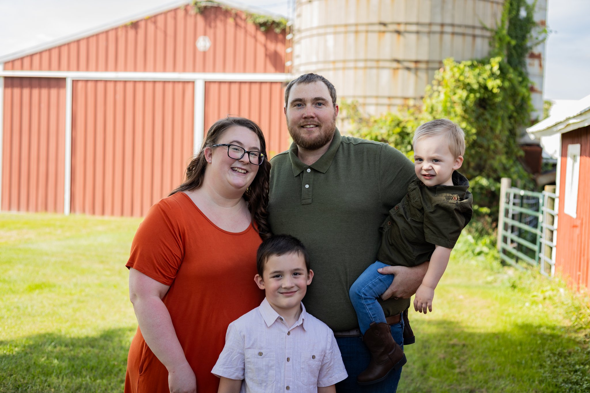 A family of four smiling together in front of a red barn