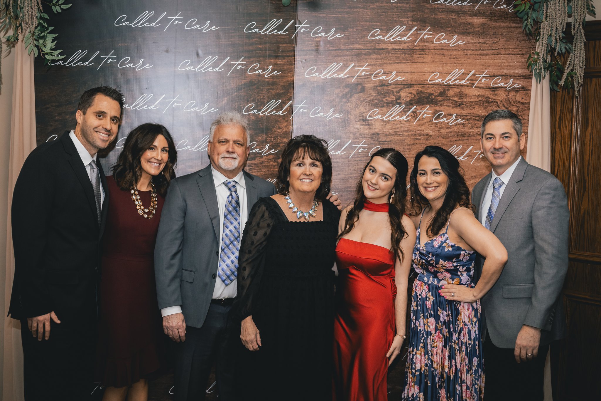 Seven formally dressed guests posing together at a Called to Care event backdrop