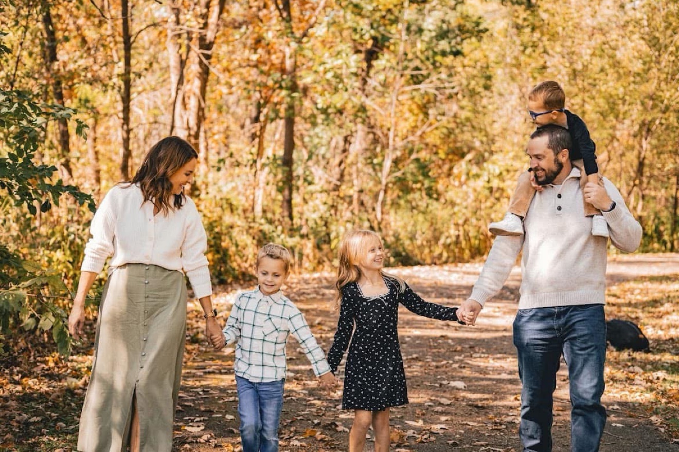 A family walking hand in hand along a wooded fall path, one child riding on dad's shoulders