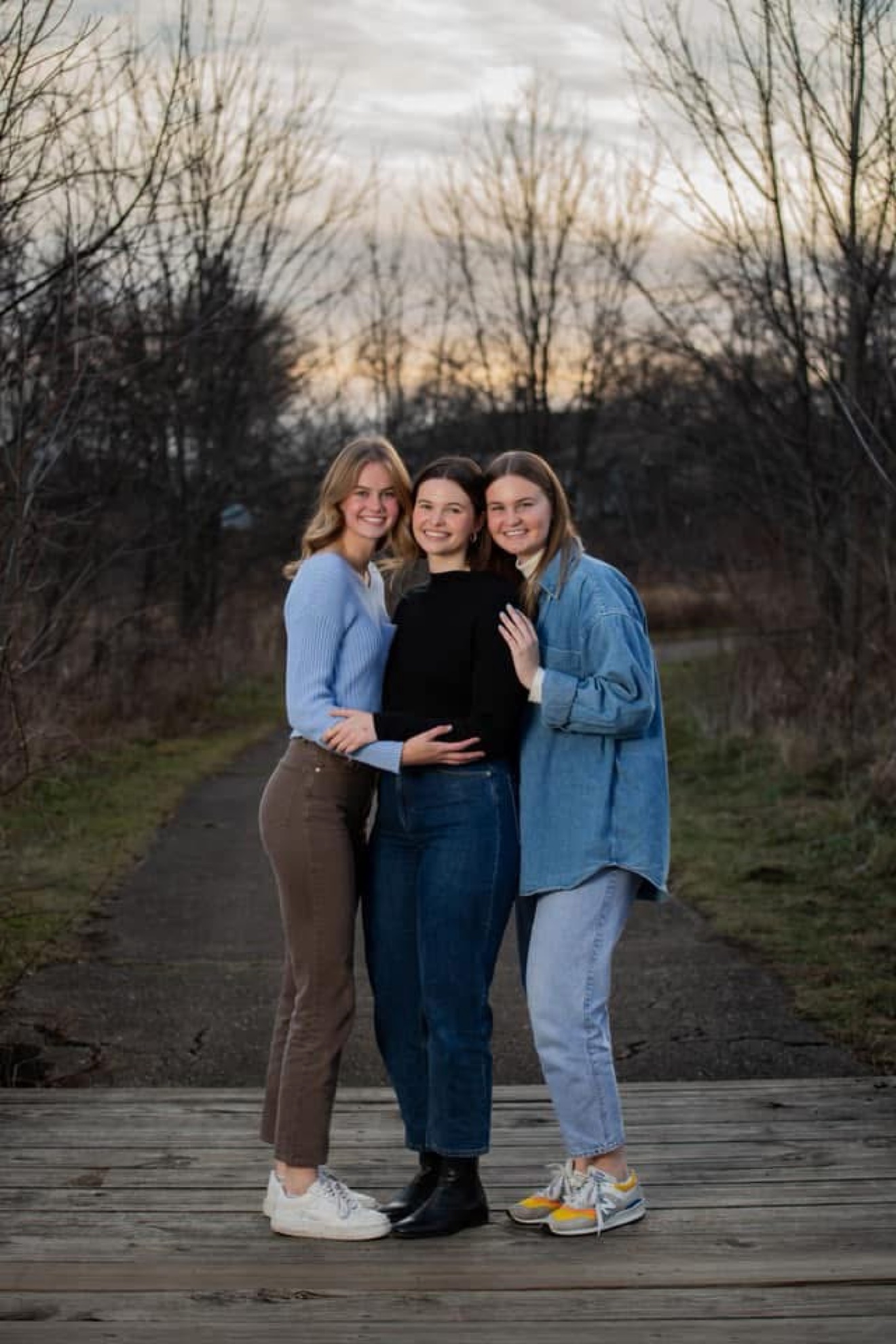 Three sisters hugging and smiling together on a wooden boardwalk at dusk