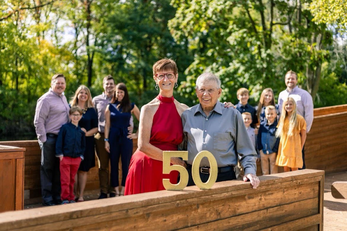 An extended family gathered behind a couple celebrating their 50th anniversary, with a gold 50 sign on a wooden rail