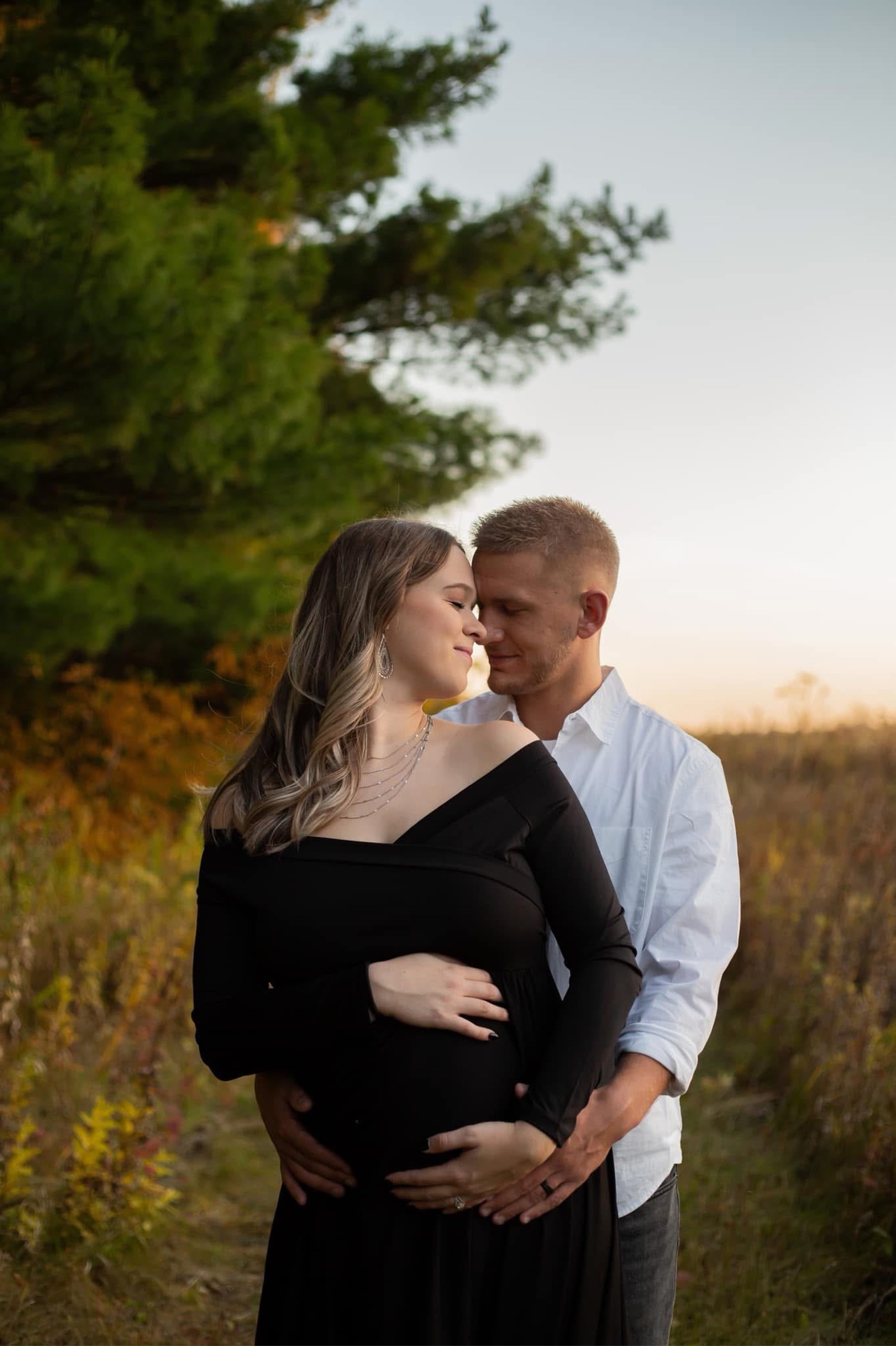 An expectant couple embracing outdoors at sunset, hands resting on her belly