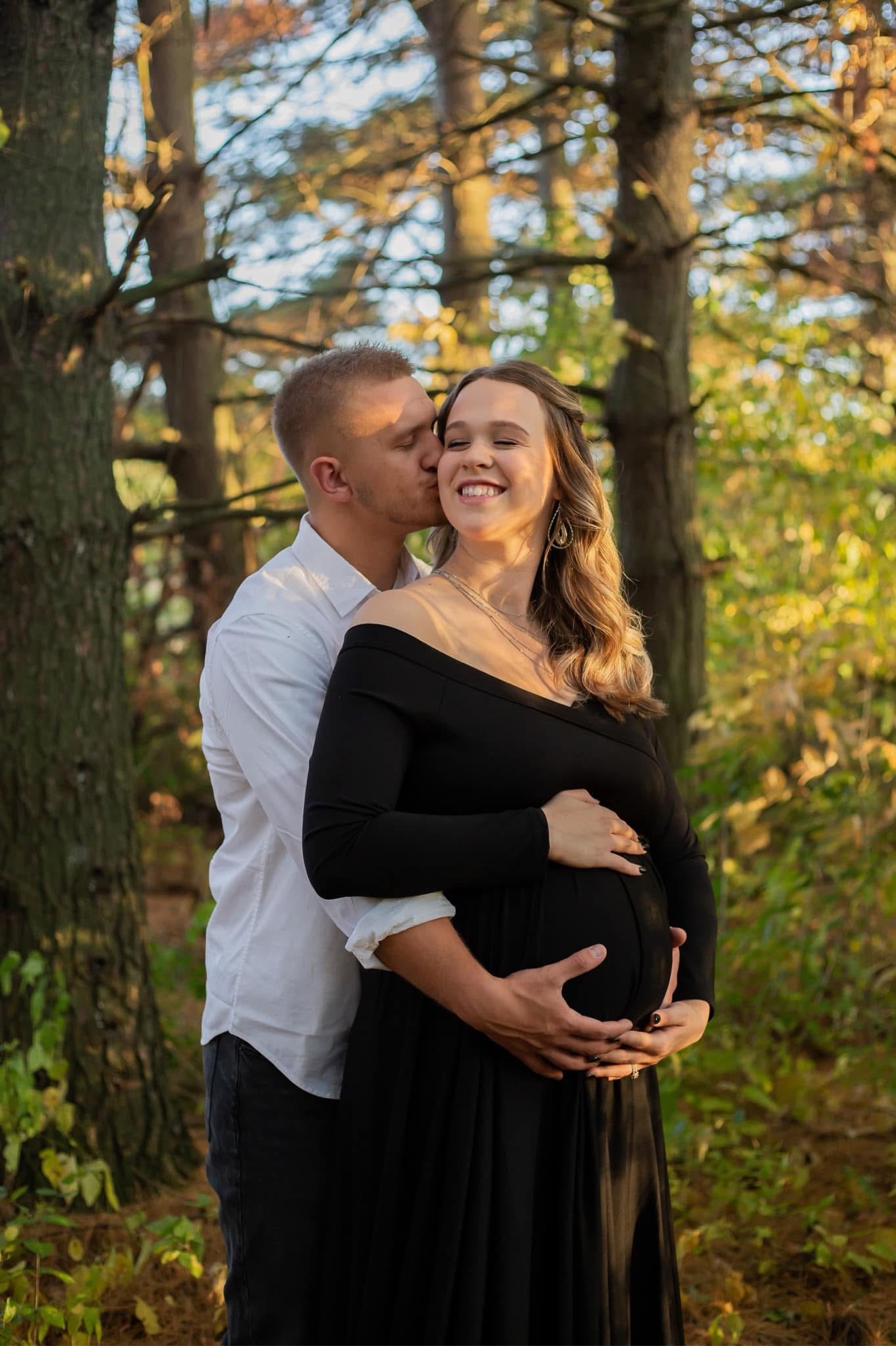 A partner kissing his expectant wife's cheek in the woods, both hands resting on her bump
