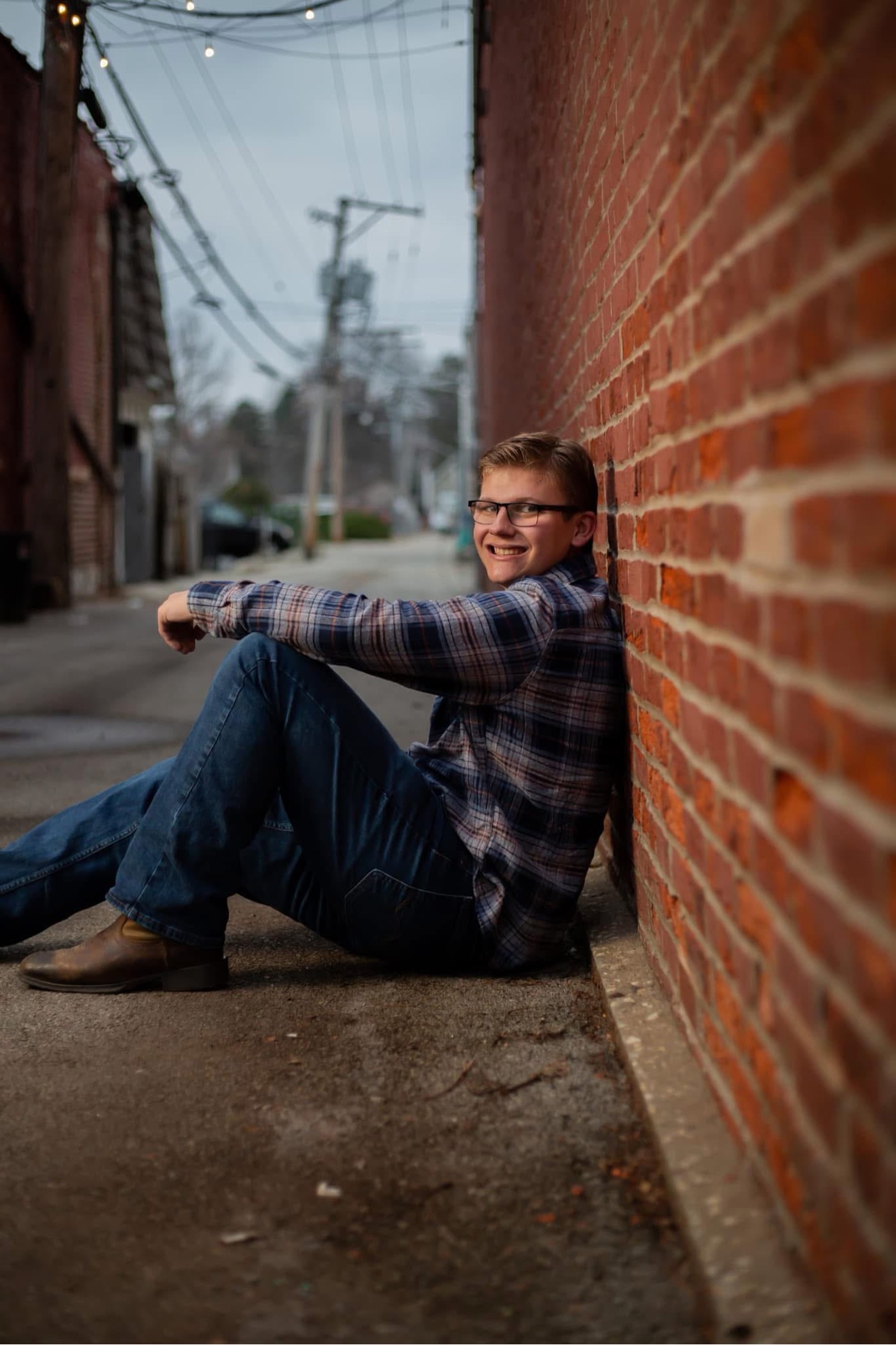 A senior portrait of a young man in a plaid shirt sitting against a red brick wall in an alley