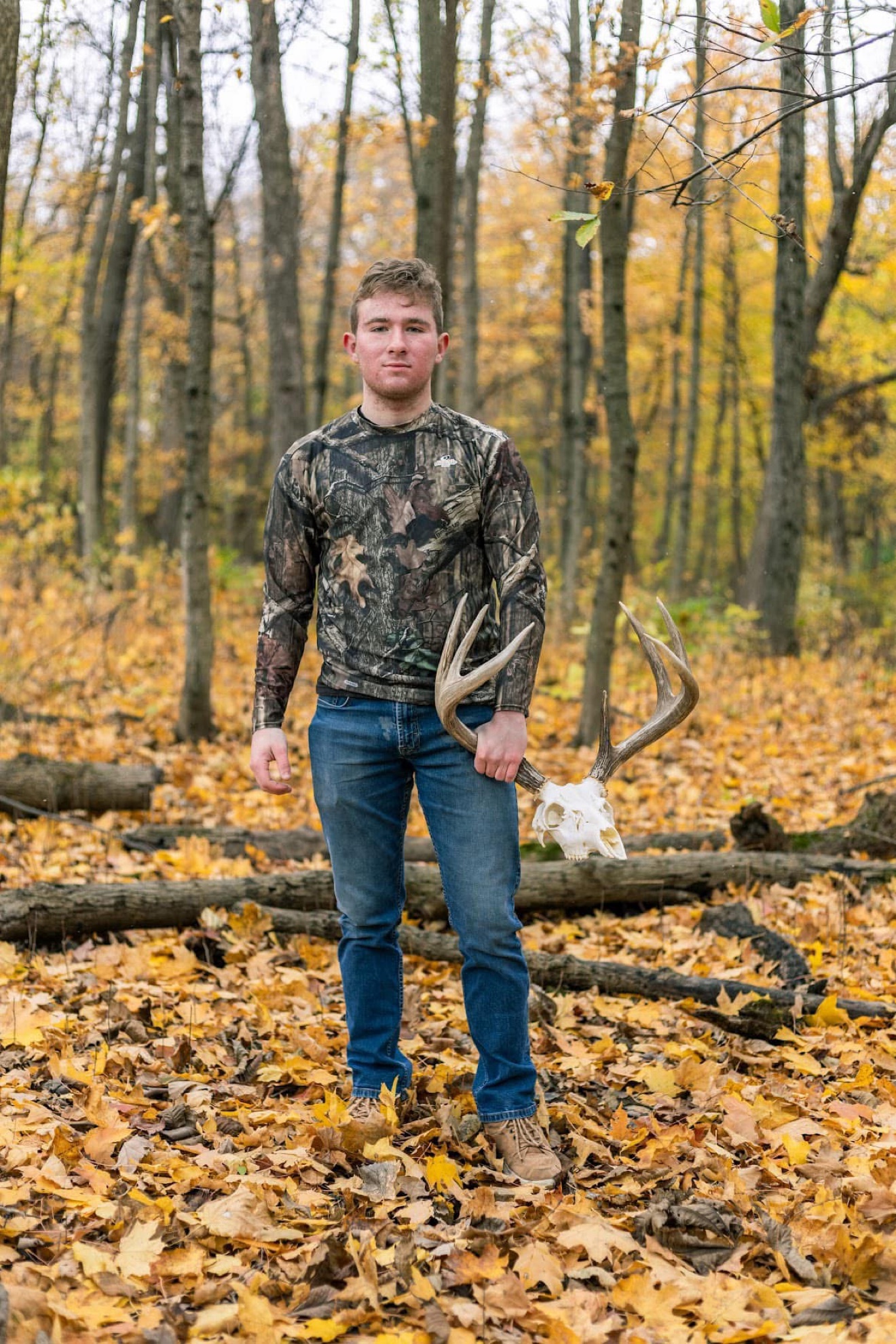 A senior portrait of a young hunter in camouflage holding a deer skull in the fall woods