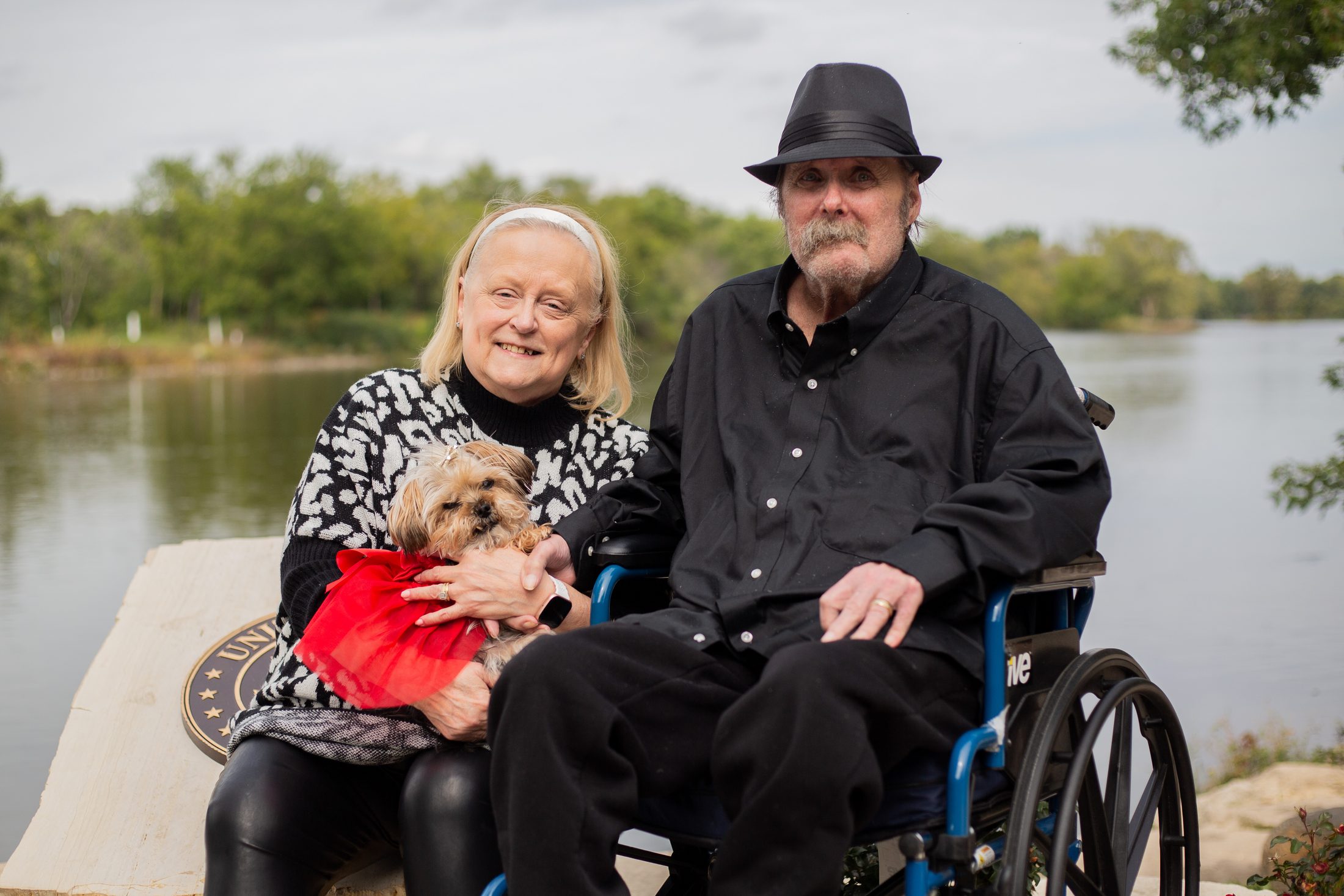 Ron and Ruth Ann, an older couple sitting by a lake, Ruth Ann holding a small dog