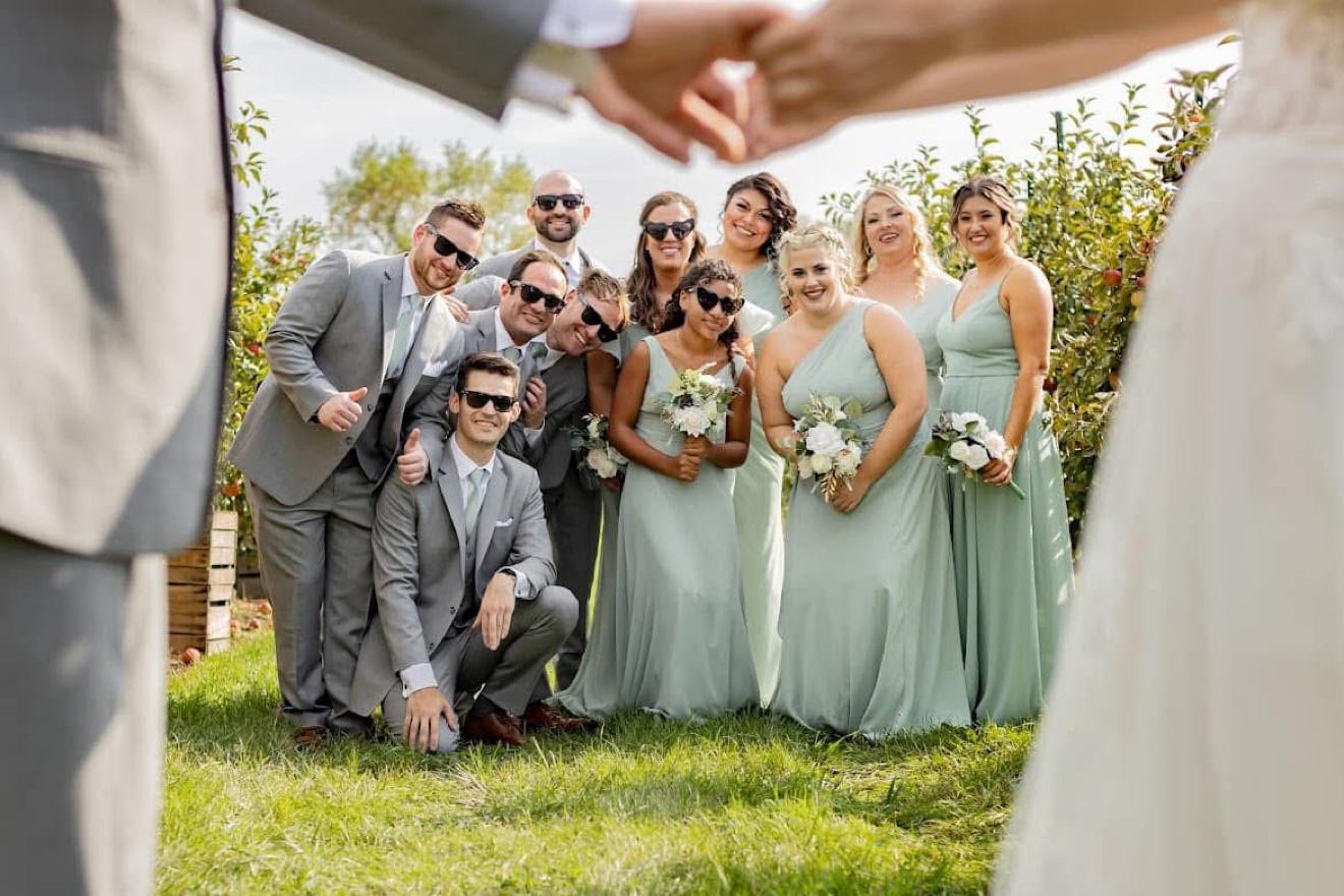 A wedding party in sage green dresses and gray suits posing in an orchard, framed by the couple's joined hands