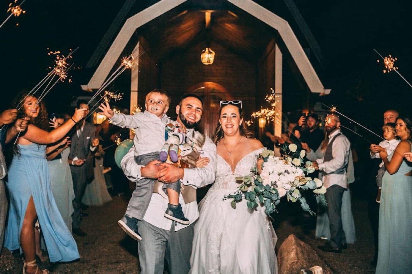 A bride and groom walking through a sparkler send-off at night, the groom carrying a young boy